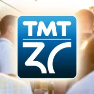 TMT-Logo zum 30-jährigen Jubiläum vor unscharfem Hintergrund mit feiernden Gästen bei Sonnenuntergang