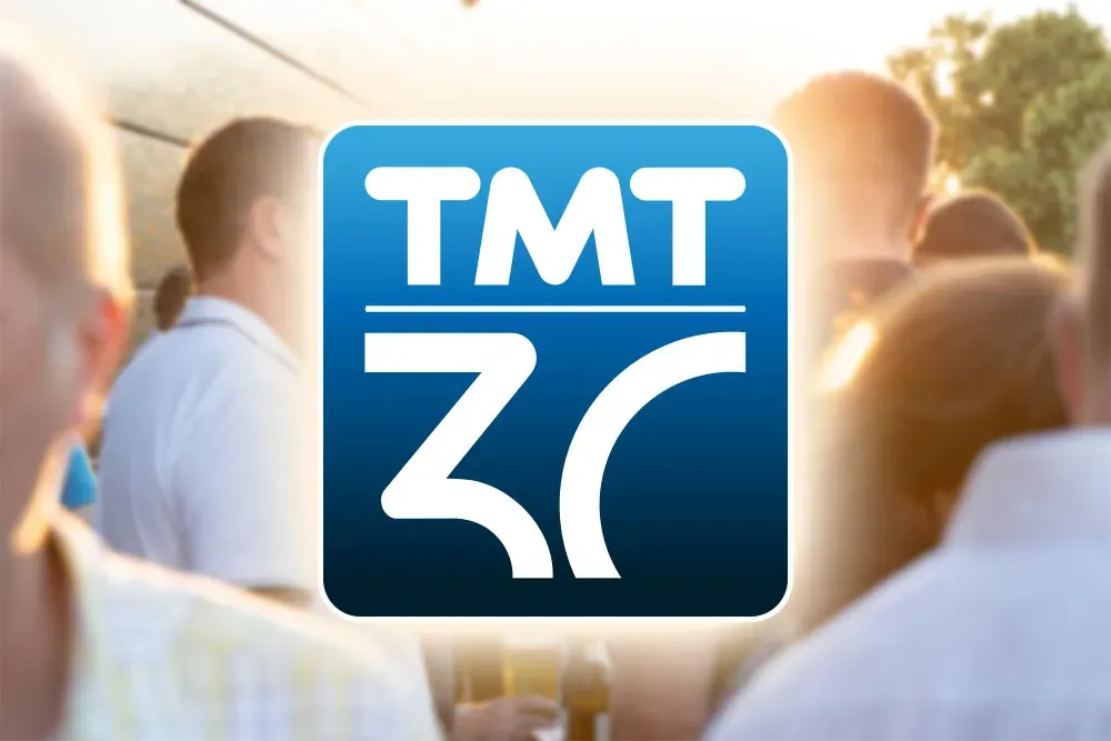 TMT-Logo zum 30-jährigen Jubiläum vor unscharfem Hintergrund mit feiernden Gästen bei Sonnenuntergang