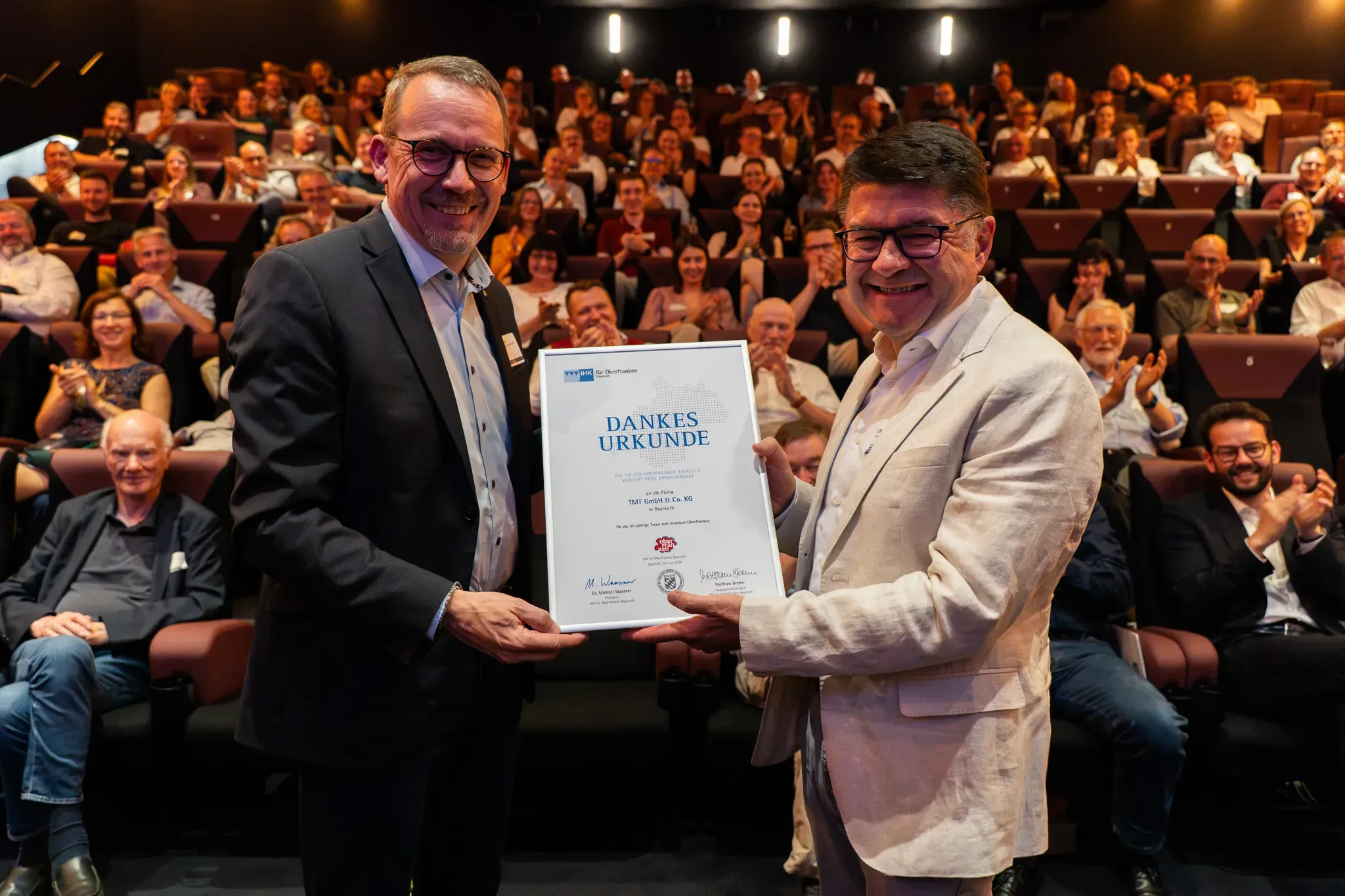 Übergabe einer Dankesurkunde auf der Bühne vor applaudierendem Publikum